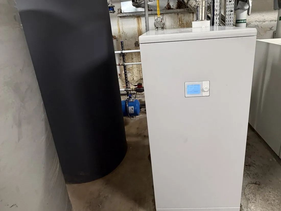 Dans une chaufferie en sous-sol, une chaudière gaz à condensation blanche au panneau de régulation bleu, à côté d’un ballon d’eau chaude gris anthracite.